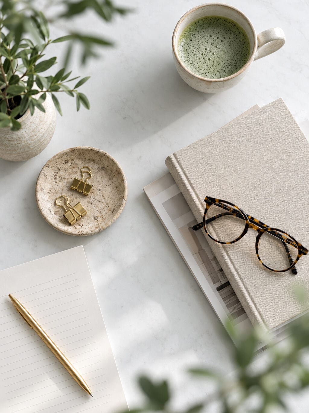 Flat lay of a cup of matcha tea, reading glasses and a gold pen on a linen notebook