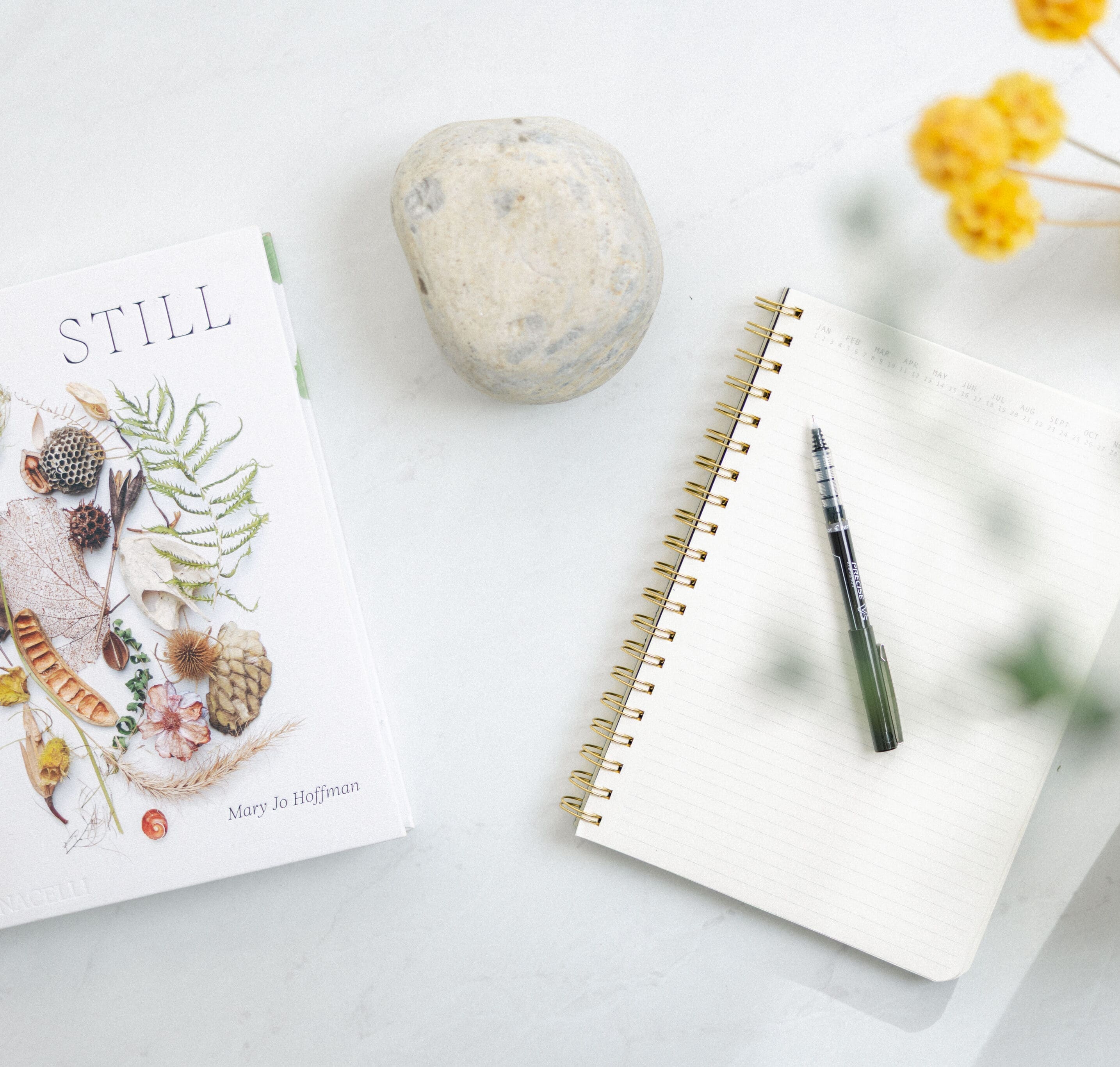 Flat lay of a spiral notebook, pen, smooth stone and yellow flowers on a white desk