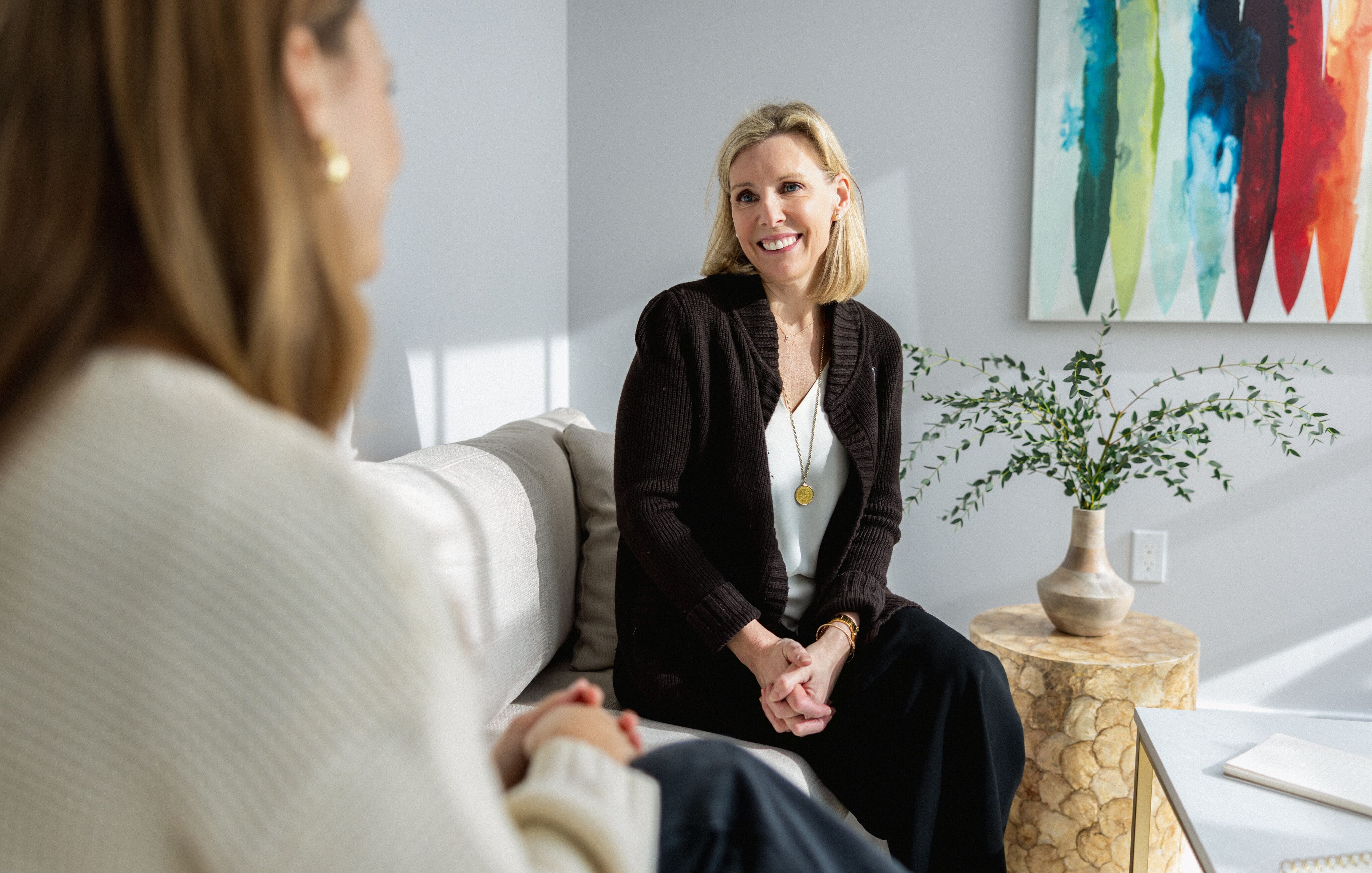 Elisabeth Tullis Schneider seated in an armchair smiling at a client, with a colorful abstract painting behind her